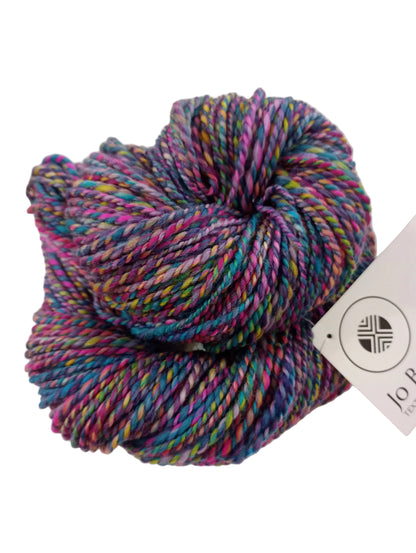 A skein of variegated yarn with purple, blue, pink, green, and yellow threads, resting on a white paper bag with a visible Jo Prudham tag.