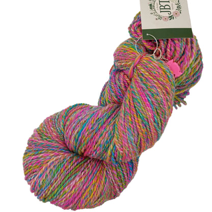 A skein of variegated yarn with pink, purple, blue, green, and yellow strands, featuring a paper brand tag and a small pink heart tag, photographed on a plain white background.